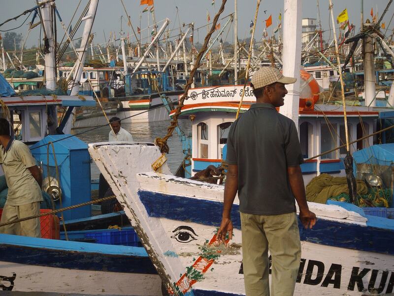 A fisherman stands beside a wooden boat with an eye painted on its bow; trawlers and flags behind.