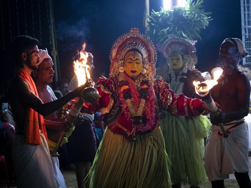 Bhuta kola performers in tender coconut-leaf skirts at night, with attendants holding oil torches.